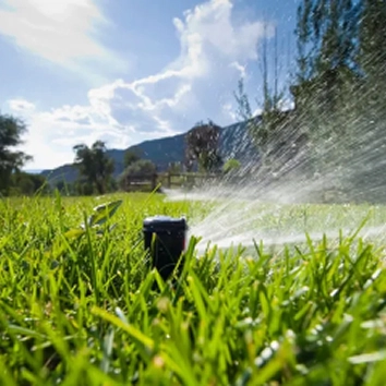 A small black sprinkler nozzle spraying water across a green grassy field on a sunny day, with hills and a fence in the background.