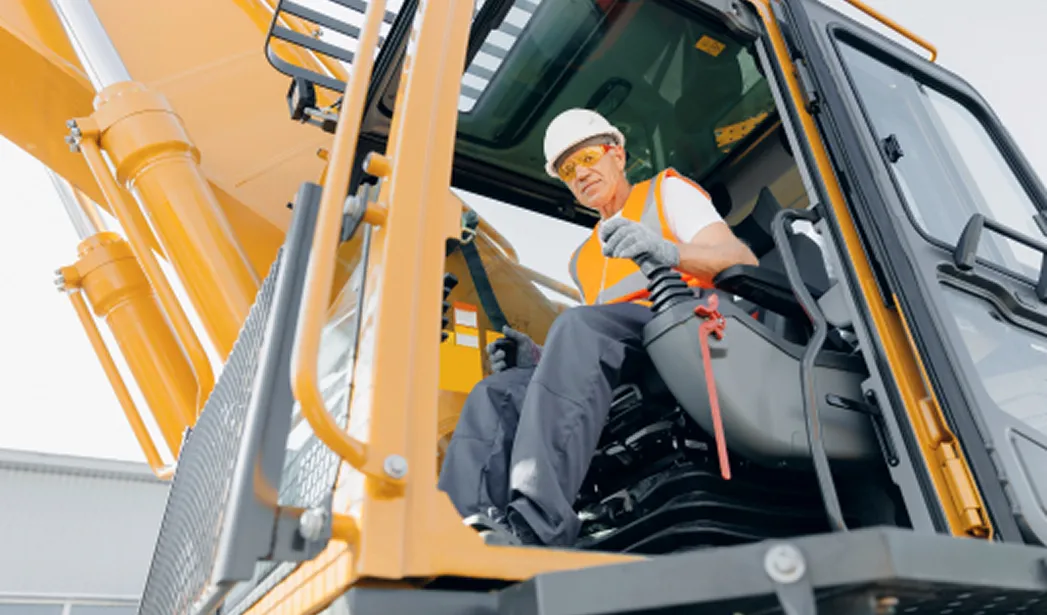 A construction worker wearing a white hard hat, orange safety vest, and protective eyewear sits inside the cab of a large yellow/orange excavator, gripping the controls. The machinery is angled, with metal arms and a grated step visible on the left.
